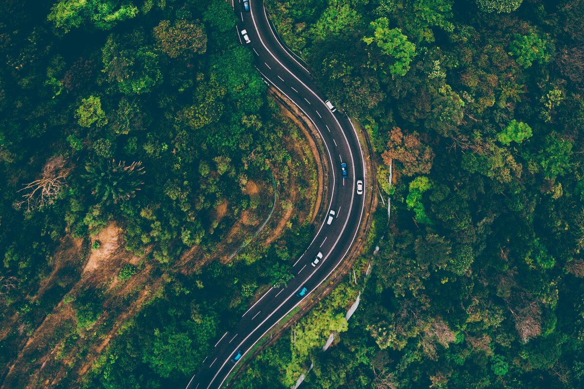 Aerial view of cars traveling along a winding two-lane road that curves through dense green forest.