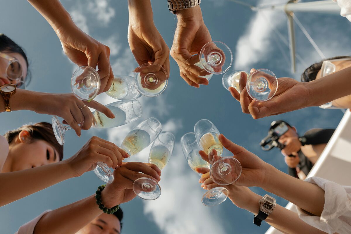 A group of people seen from below raising and clinking glasses of sparkling wine against a blue sky.