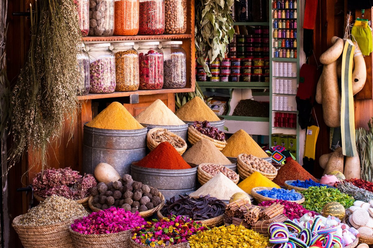 A market stall overflowing with cones of colorful spices, baskets of dried flowers and goods, and shelves of glass jars.